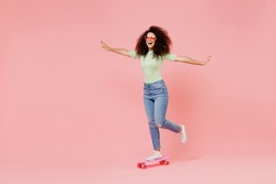 Full size body length happy fun excited bright young curly latin woman 20s wears casual clothes sunglasses skate on board spreading hands isolated on plain pastel light pink background studio portrait