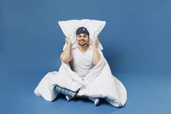 Full size body length happy young man in pajamas jam sleep mask rest relax at home cover ears with pillow sit wrap cover blanket duvet isolated on dark blue background Good mood night bedtime concept
