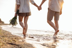 Cropped close up back view young couple two family man woman in white clothes hold flowers bouquet hand walk in water waves at sunrise over sea beach ocean outdoor seaside in summer day sunset evening