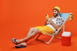 Young fun happy tourist man in beach shirt hat lie on deckchair near fridge hold use mobile cell phone isolated on plain orange background studio portrait. Summer vacation sea rest sun tan concept.
