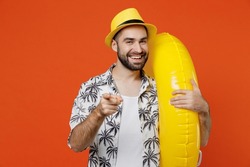 Young smiling tourist man 20s wear beach shirt hat hold inflatable ring point index finger camera on you isolated on plain orange background studio portrait. Summer vacation sea rest sun tan concept