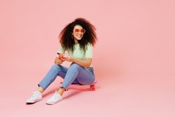 Full size body length young curly latin woman 20s wears casual clothes sunglasses sit on skateboard look aside hold use mobile cell phone isolated on plain pastel light pink background studio portrait