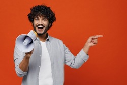 Young bearded Indian man 20s wears blue shirt hold scream in megaphone announces discounts sale Hurry up point on workspace area copy space mock up isolated on plain orange background studio portrait