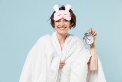 Young smiling happy woman in pajamas jam sleep eye mask rest relax at home wrap covered under blanket duvet hold clock alarm isolated on pastel blue background studio Good mood night bedtime concept