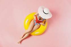 Top view full size young woman in striped swimsuit lies on inflatable rubber ring pool read book cover face with hat isolated on plain pastel pink background. Summer vacation sea rest sun tan concept