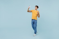 Full body young man wear yellow t-shirt doing selfie shot on mobile cell phone post photo on social network show v-sign isolated on plain pastel light blue background studio. People lifestyle concept