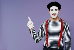 Happy charismatic fun young mime man with white face mask wears striped shirt beret pointing aside on workspace area copy space mock up isolated on plain pastel light violet background studio portrait