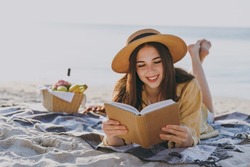Full size young happy traveler tourist woman 20s in straw hat shirt summer clothes read book lying on plaid have picnic outdoor on sea sand beach background People vacation lifestyle journey concept