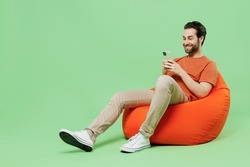 Full size young man 20s wear casual orange t-shirt sit in bag chair hold in hand use mobile cell phone isolated on plain pastel light green color background studio portrait. People lifestyle concept
