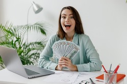 Young amazed employee business woman in blue shirt holding fan of cash money in dollar banknotes sit work at workplace white desk with laptop pc computer at office indoors. Achievement career concept