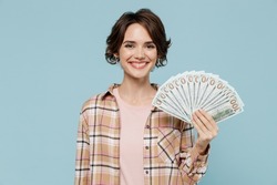 Young smiling happy woman 20s wearing casual brown shirt holding fan of cash money in dollar banknotes isolated on pastel plain light blue color background studio portrait. People lifestyle concept