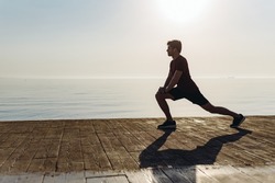 Full size side view young strong sporty athletic toned fit sportsman man in sports clothes do squates warm up training at sunrise sun dawn over sea beach outdoor on pier seaside in summer day morning