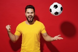 Exultant fun young bearded man football fan in yellow t-shirt cheer up support favorite team tossed soccer ball celebrate clenching fists say yes isolated on plain dark red background studio portrait