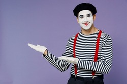 Happy smiling young mime man with white face mask wears striped shirt beret pointing aside on empty palm with copy space place mock up isolated on plain pastel light violet background studio portrait
