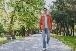 Full length young man 20s he wearing orange jacket blue t-shirt walking look camera rest relax in spring green city park go down alley sunshine lawn outdoors on nature. Urban lifestyle leisure concept