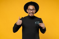 Young happy smiling successful american african man 20s in stylish black shirt hat eyeglasses holding credit bank card show thumb up like gesture isolated on yellow orange background studio portrait.