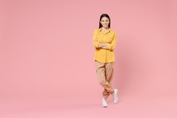 Full length of young smiling pretty happy brunette positive attractive beautiful latin woman 20s in yellow casual shirt hold hands crossed folded isolated on pastel pink background studio portrait
