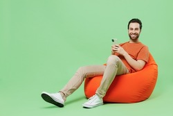 Full length young man 20s wear casual orange t-shirt sit in bag chair hold in hand use mobile cell phone isolated on plain pastel light green color background studio portrait. People lifestyle concept