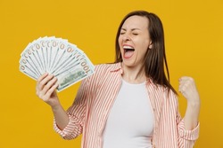 Young happy exultant overjoyed excited woman she 30s wears striped shirt white t-shirt hold fan of cash money in dollar banknotes do winner gesture isolated on plain yellow background studio portrait.