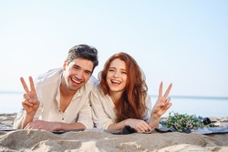 Enamored happy smiling young couple two family man woman in white clothes lying on picnic plaid show v-sign rest relax together at sunrise over sea beach ocean outdoor seaside in summer day sunset.