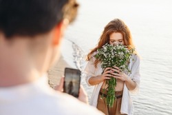 Happy young couple family man woman in white clothes sniff flowers bouquet take photo on mobile cell phone rest together at sunrise over sea beach ocean outdoor seaside in summer day sunset evening