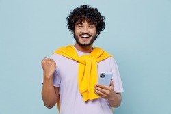 Happy young bearded Indian man 20s wears white t-shirt hold in hand use mobile cell phone doing winner gesture celebrate clenching fists isolated on plain pastel light blue background studio portrait