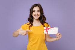 Smiling attractive young brunette woman 20s wearing basic yellow t-shirt posing hold in hands pointing index finger on gift certificate isolated on pastel violet colour background, studio portrait