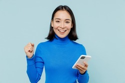 Fun young woman of Asian ethnicity 20s years old wears blue shirt hold in hand use mobile cell phone doing winner gesture clenching fists isolated on plain pastel light blue background studio portrait