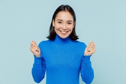 Overjoyed excited exultant young woman of Asian ethnicity 20s years old wears blue shirt doing winner gesture celebrate clenching fists isolated on plain pastel light blue background studio portrait