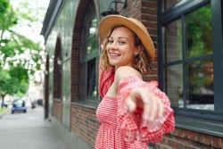 Young smiling fun happy woman in pink dress hat look camera stretch hand say follow me walk in city center standing outdoor near town brick old building. People urban summer time lifestyle concept.