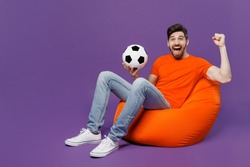 Full body young fan man he wear orange t-shirt cheer up support football sport team hold soccer ball watch tv live stream sit in bag chair do winner gesture isolated on plain dark purple background