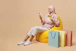 Full body young arabian asian muslim woman she wear abaya hijab sit in bag chair hold mobile phone credit card shopping isolated on plain light beige background. People uae islam religious concept.