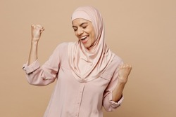 Young arabian asian muslim woman she wear abaya hijab pink clothes do winner gesture celebrate clench fist isolated on plain pastel beige background. People uae middle eastern islam religious concept