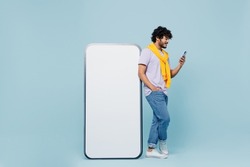 Full size young bearded Indian man 20s wears white t-shirt hold in hand stand near big mobile cell phone with blank screen workspace area isolated on plain pastel light blue background studio portrait