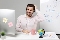 Employee puzzled troubled operator business man in set microphone headset for helpline assistance sit work at call center office desk with pc computer scratch head isolated on grey background studio
