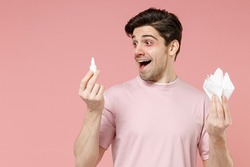 Fun shocked sick unhealthy ill allergic man has red eyes runny stuffy sore nose suffer from allergy symptoms hay fever hold paper napkin use nasal drops isolated on pastel pink color background studio