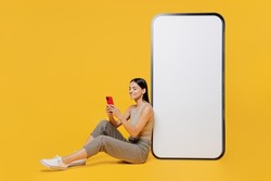 Full size smiling happy young latin woman she wear tank shirt sit near big huge blank screen mobile cell phone with workspace copy space mockup area use smartphone isolated on plain yellow backround