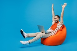 Full body young excited fun cool man 20s in orange striped t-shirt sit in bag chair hold use work on laptop pc computer with overstretched hands legs isolated on plain blue background studio portrait.