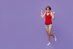 Full body young smiling woman 20s she wear red tank shirt eyeglasses doing winner gesture celebrate clenching fists say yes isolated on plain purple background studio portrait People lifestyle concept