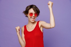 Young overjoyed happy woman 20s she wear red tank shirt eyeglasses doing winner gesture celebrate clenching fists say yes isolated on plain purple background studio portrait. People lifestyle concept