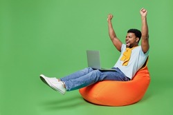 Full body excited young man of African American ethnicity 20s in blue t-shirt sit in bag chair hold use work on laptop pc computer do winner gesture isolated on plain green background studio portrait.
