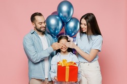 Young parents mom dad with child kid daughter teen girl in blue clothes celebrating birthday party hold bunch air inflated balloons red present box cover eyes isolated on plain pastel pink background.
