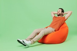 Full body smiling happy fun cool young man 20s wear casual orange t-shirt sit in bag chair hold hands behind neck isolated on plain pastel light green color background studio People lifestyle concept