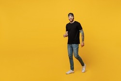 Full body young happy bearded tattooed man 20s he wears casual black t-shirt cap walking going strolling look camera isolated on plain yellow wall background studio portrait. People lifestyle concept
