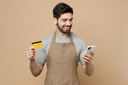 Young man barista barman employee wear brown apron work in coffee shop using mobile cell phone credit bank card shopping online isolated on plain light beige background Small business startup concept