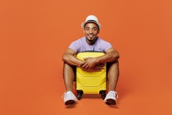 Full body traveler fun black man wear purple t-shirt hat sit hold suitcase isolated on plain orange color background. Tourist travel abroad in spare time rest getaway. Air flight trip journey concept