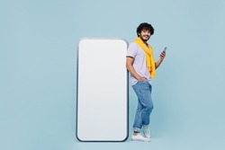Full size fancy young bearded Indian man 20s wears white t-shirt hold stand near big mobile cell phone with blank screen workspace area isolated on plain pastel light blue background studio portrait