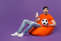Full size young fan man he wear orange t-shirt cheer up support football sport team hold soccer ball mobile cell phone watch tv live stream sit in bag chair isolated on plain dark purple background