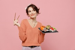 Young minded pensive thoughtful fun woman in sweater hold in hand makizushi sushi roll served on black plate traditional japanese food look aside chopsticks isolated on plain pastel pink background.