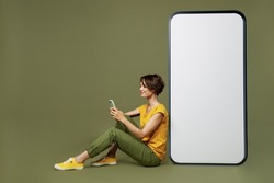 Full body young woman she 20s wear yellow t-shirt sit near big huge blank screen mobile cell phone with workspace copy space mockup area use smartphone isolated on plain olive green khaki background
